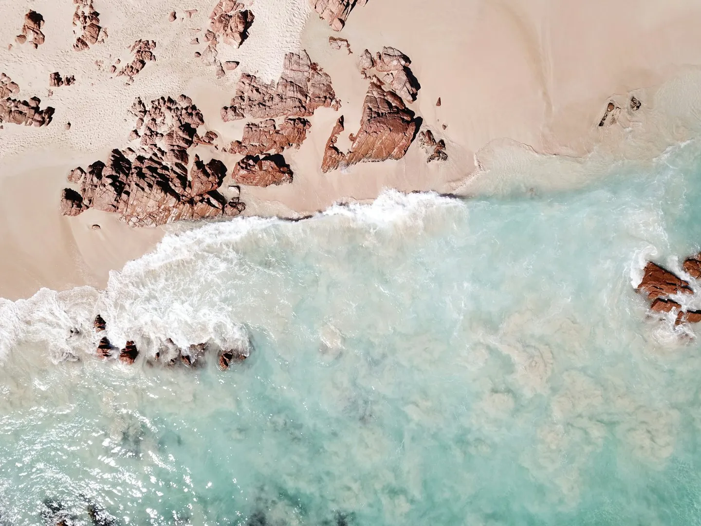 Top-down aerial of turquoise surf meeting rust-coloured coastal rocks